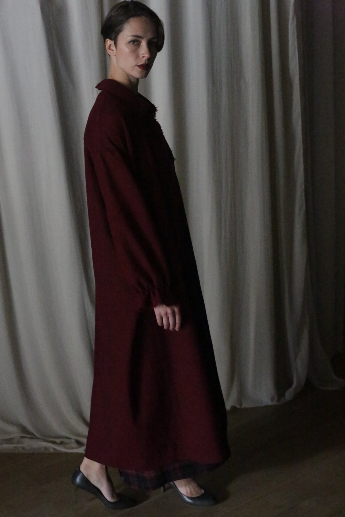 A person stands sideways in the Dress Coat | Japanese Wool Gauze Wine and heels, looking at the camera. Neutral curtains and soft, dim lighting create a subtle backdrop.