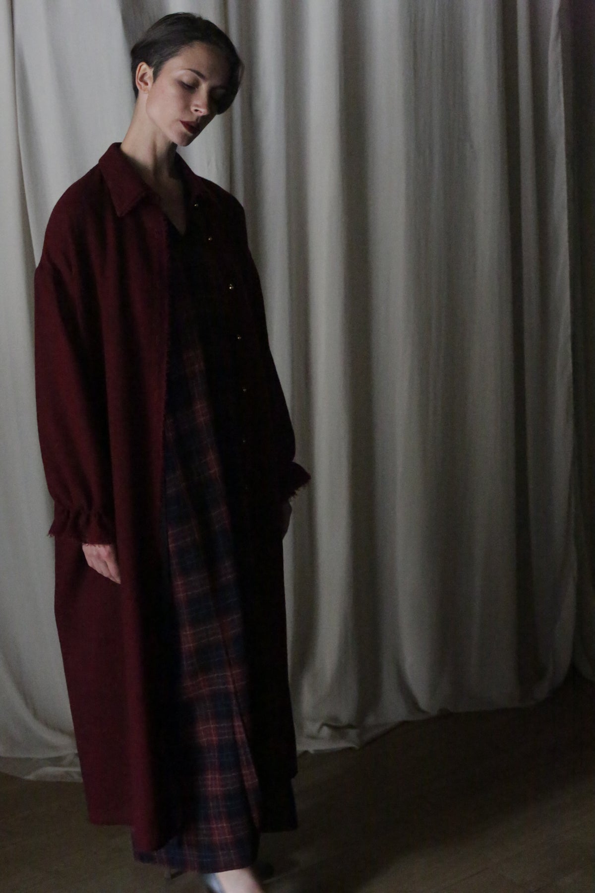 A person with short dark hair stands indoors against white curtains, looking down calmly while wearing the Dress Coat | Japanese Wool Gauze Wine over a plaid dress.