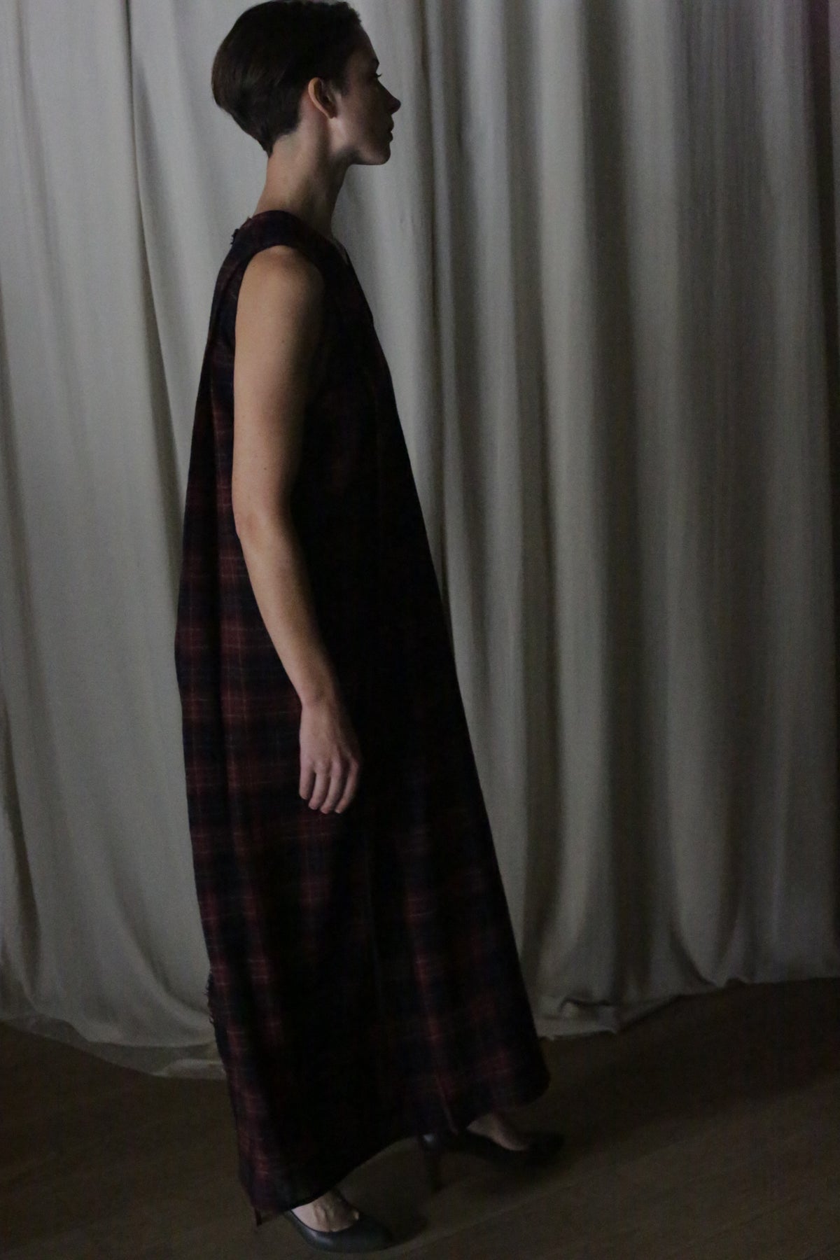A person with short dark hair, wearing The Winter Dress in Japanese Wool Gauze Wine Plaid and dark shoes, stands in profile before light-colored curtains.