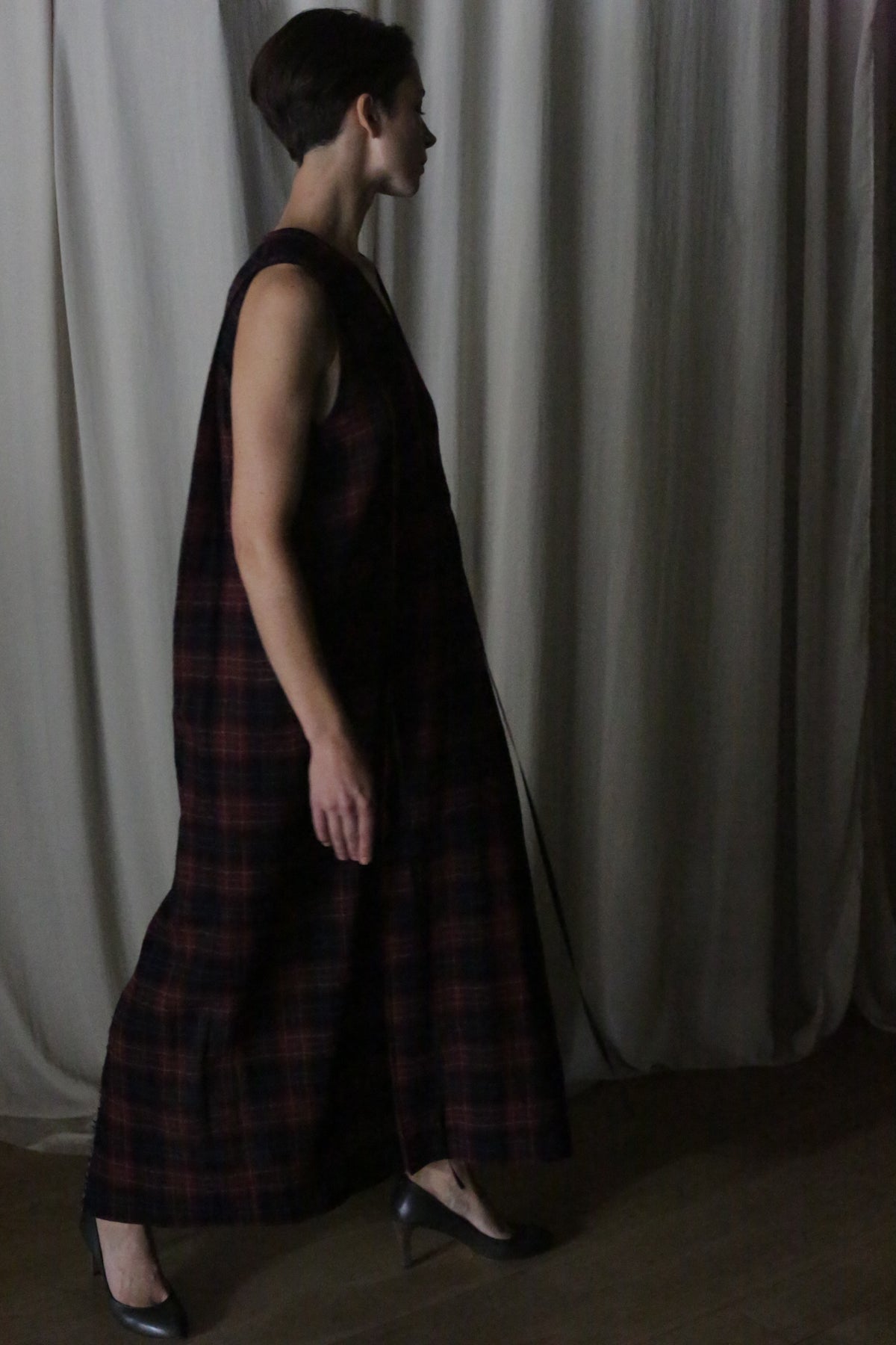 A person with short hair, wearing The Winter Dress | Japanese Wool Gauze Wine Plaid and black heels, walks indoors before light curtains as dim lighting creates soft shadows.