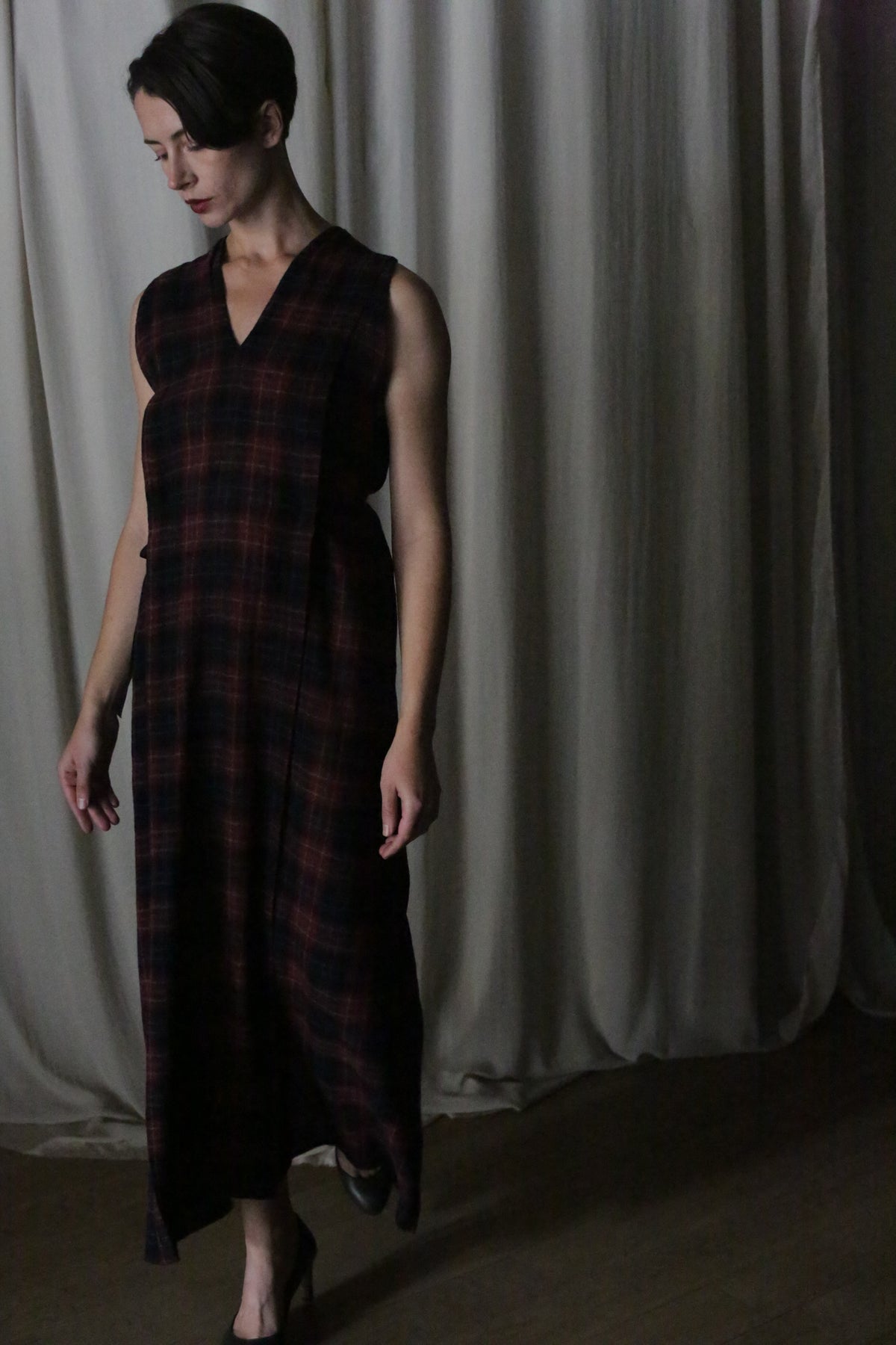 A person with short dark hair and a neutral expression stands indoors on a wooden floor in front of pale curtains, wearing The Winter Dress | Japanese Wool Gauze Wine Plaid.