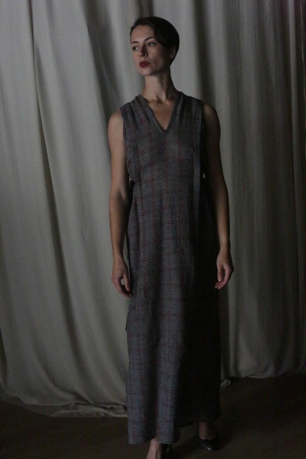 A person with short dark hair stands before light curtains, wearing The Winter Dress in Japanese Wool Gauze Grey Plaid—an ankle-length, sleeveless winter dress—and gazes slightly to the side with a neutral expression.
