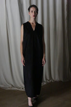 Load image into Gallery viewer, In a dimly lit room, someone dons &quot;The Winter Dress | Cashmere Black,&quot; a long, sleeveless black cashmere dress. They have short, dark hair and wear a serious expression as they stand in front of a softly lit curtain.