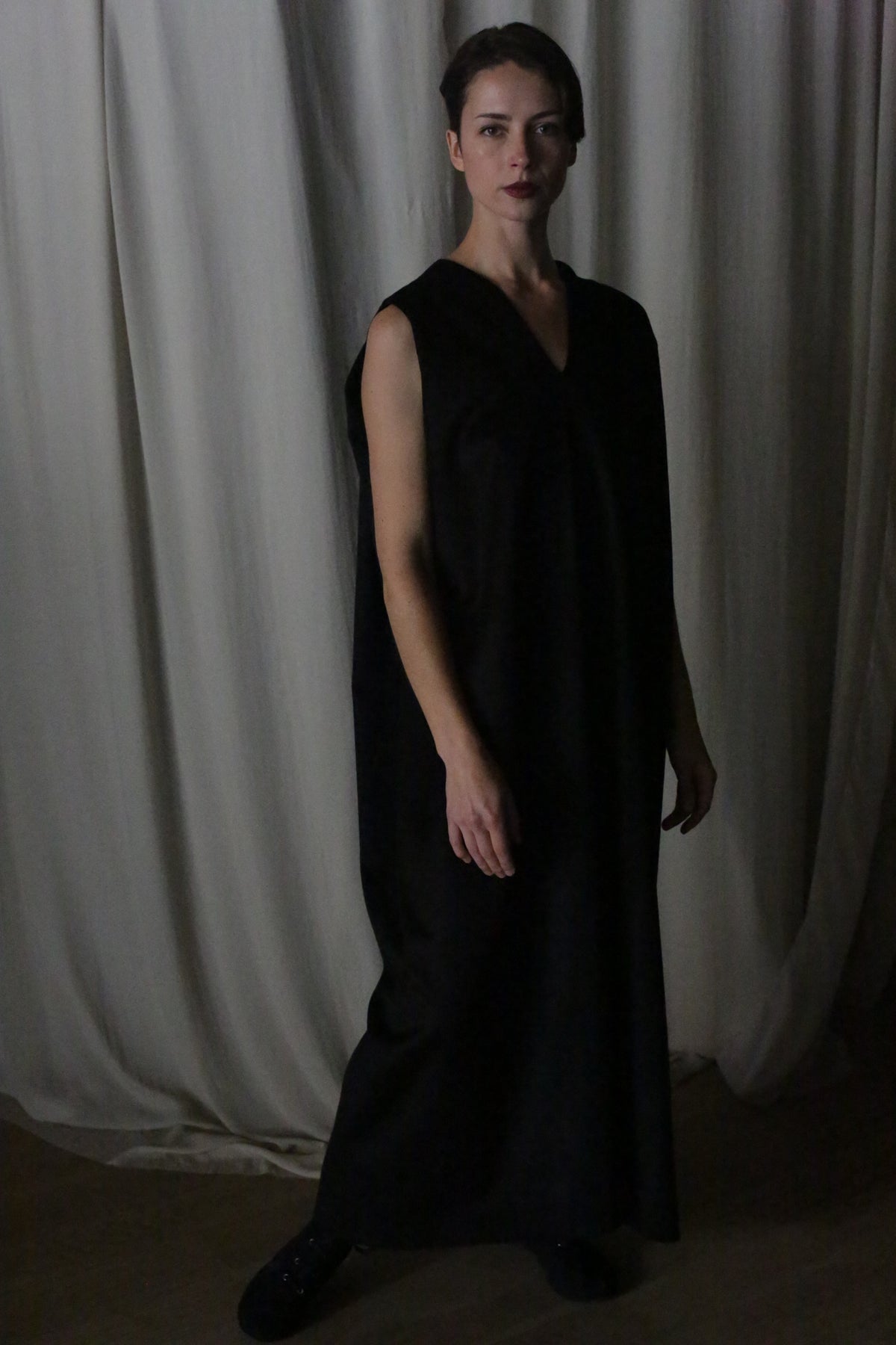 A person stands in a dimly lit room wearing The Winter Dress in black cashmere. The background features draped white curtains, adding to the serene atmosphere.