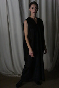 A person stands in a dimly lit room wearing The Winter Dress in black cashmere. The background features draped white curtains, adding to the serene atmosphere.