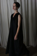 Load image into Gallery viewer, A person wearing The Winter Dress in cashmere black, which is long and sleeveless, stands in front of a light gray curtain, looking off to the side with a neutral expression.