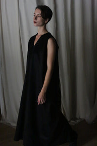 A person wearing The Winter Dress in cashmere black, which is long and sleeveless, stands in front of a light gray curtain, looking off to the side with a neutral expression.