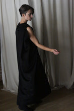 Load image into Gallery viewer, A person with short hair is wearing The Winter Dress | Cashmere Black. They are positioned on a wooden floor in front of sheer white curtains, turned slightly to the side, with one arm gracefully extended.