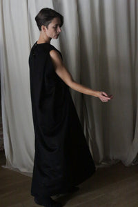 A person with short hair is wearing The Winter Dress | Cashmere Black. They are positioned on a wooden floor in front of sheer white curtains, turned slightly to the side, with one arm gracefully extended.