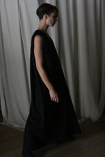 Load image into Gallery viewer, A person wearing &quot;The Winter Dress | Cashmere Black,&quot; a long and flowing design, stands in profile against a backdrop of light-colored curtains. The dim lighting creates a dramatic silhouette, emphasizing the elegance of this handmade winter dress.