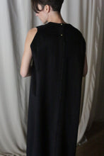 Load image into Gallery viewer, A person stands facing away from the camera in The Winter Dress | Cashmere Black, its elegant silhouette softly contrasting with the light-colored curtains.