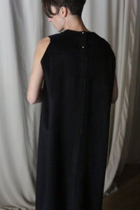 A person stands facing away from the camera in The Winter Dress | Cashmere Black, its elegant silhouette softly contrasting with the light-colored curtains.