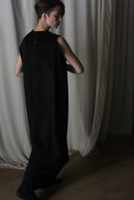 Load image into Gallery viewer, Standing in front of white curtains, a person with short dark hair showcases &quot;The Winter Dress | Cashmere Black,&quot; a long, sleeveless cashmere creation. In the dimly lit room, they glance down and to the side, exuding the cozy elegance that defines this winter piece.