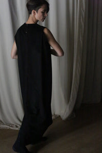 Standing in front of white curtains, a person with short dark hair showcases "The Winter Dress | Cashmere Black," a long, sleeveless cashmere creation. In the dimly lit room, they glance down and to the side, exuding the cozy elegance that defines this winter piece.