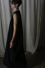 Load image into Gallery viewer, A person wearing &quot;The Winter Dress | Cashmere Black&quot; stands sideways in a dimly lit room. The light-colored draped curtains behind them enhance the elegance of their winter ensemble.