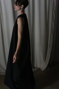 A person wearing "The Winter Dress | Cashmere Black" stands sideways in a dimly lit room. The light-colored draped curtains behind them enhance the elegance of their winter ensemble.