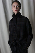 In a moody and elegant setting, an individual stands against a draped curtain, wearing the Simple Shirt in Japanese Wool Gauze Forest Plaid with a high collar and removable black ribbon tie, complemented by dark pants.
