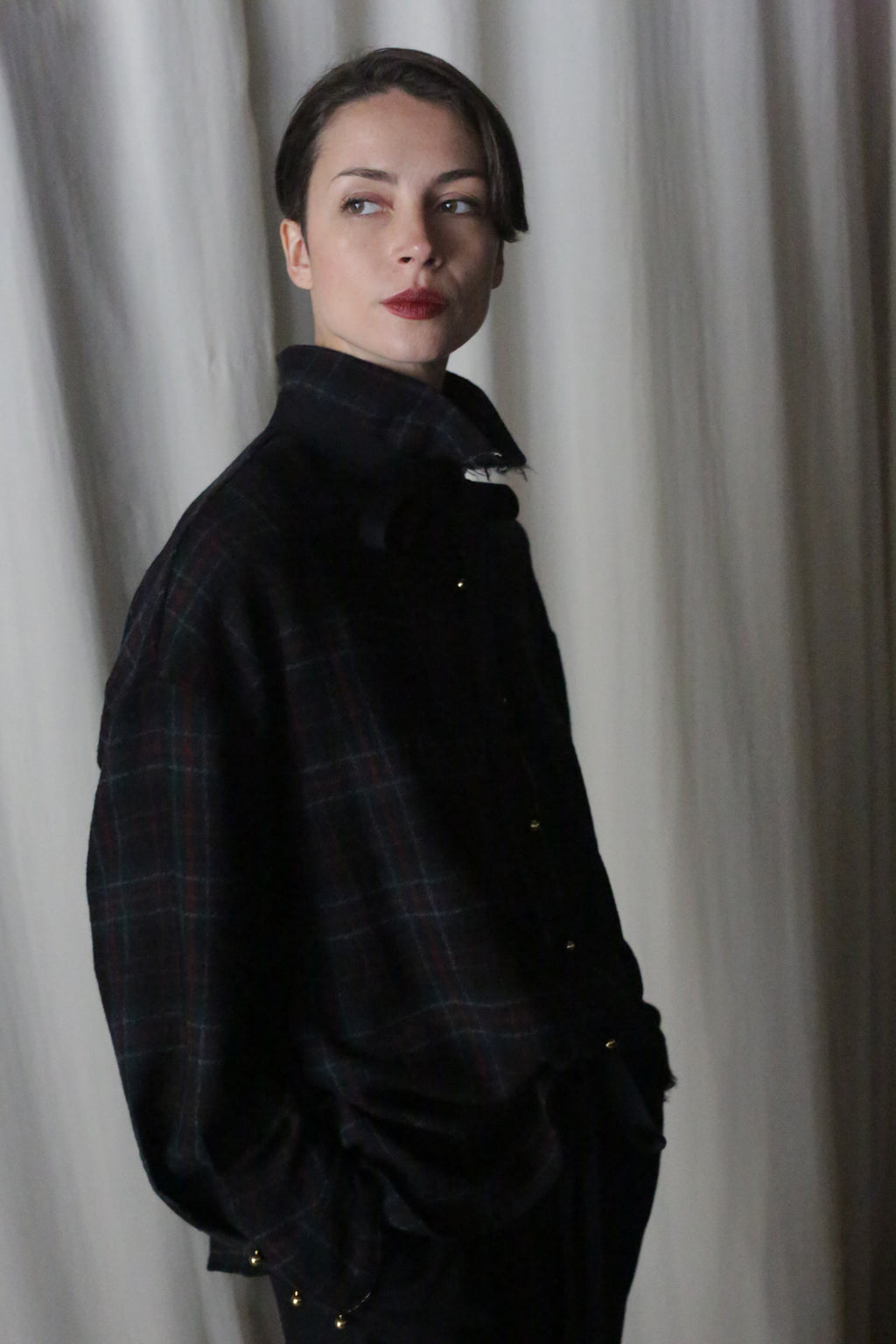 Wearing a Simple Shirt in Japanese Wool Gauze Forest Plaid, a person with short hair and red lipstick stands before a light curtain, gazing sideways. Soft lighting casts a shadow behind, highlighting the chic flair of their outfit.