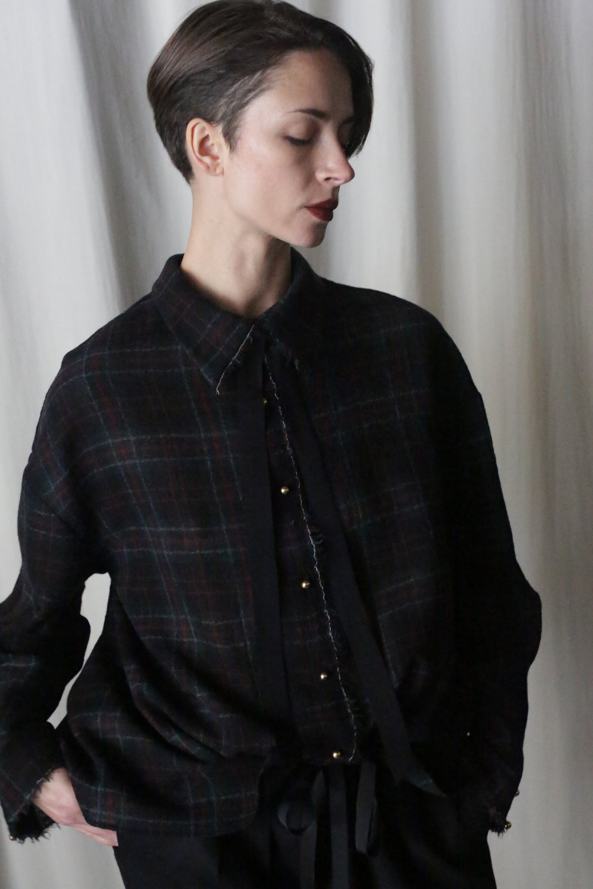 A person with short hair and closed eyes stands against a white curtain, wearing the Simple Shirt in Japanese Wool Gauze Forest Plaid, paired with a black tie and pants. Their hands are in pockets, creating a soft, introspective mood in the lighting.