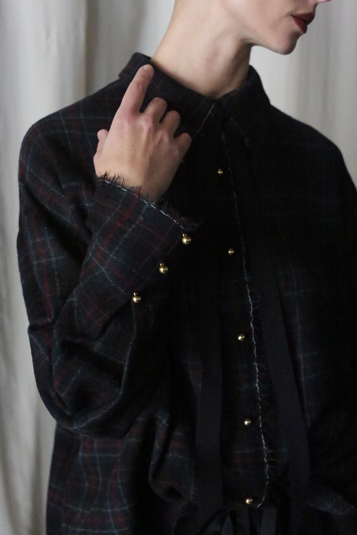 A person, wearing the Simple Shirt in Japanese Wool Gauze Forest Plaid with gold button sleeve details and a removable black ribbon tie, holds the collar. The frayed-edge fabric's festive plaids stand out against a softly lit neutral background.