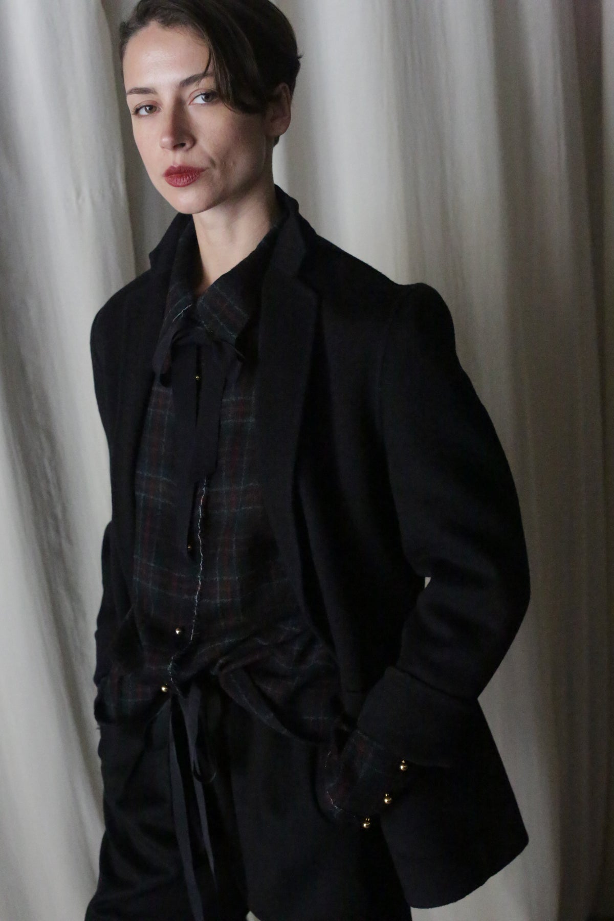 A person with short dark hair wears a Simple Shirt in Japanese Wool Gauze Forest Plaid and a dark blazer, standing before a white curtain. They have a neutral expression, hands tucked in pockets.