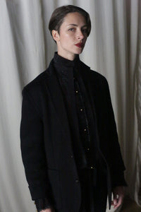 A person with short hair, dressed in the Unisex Blazer Double-Face Cashmere in charcoal, stands confidently against a backdrop of white curtains. The lighting casts a dramatic shadow, accentuating their poised expression.