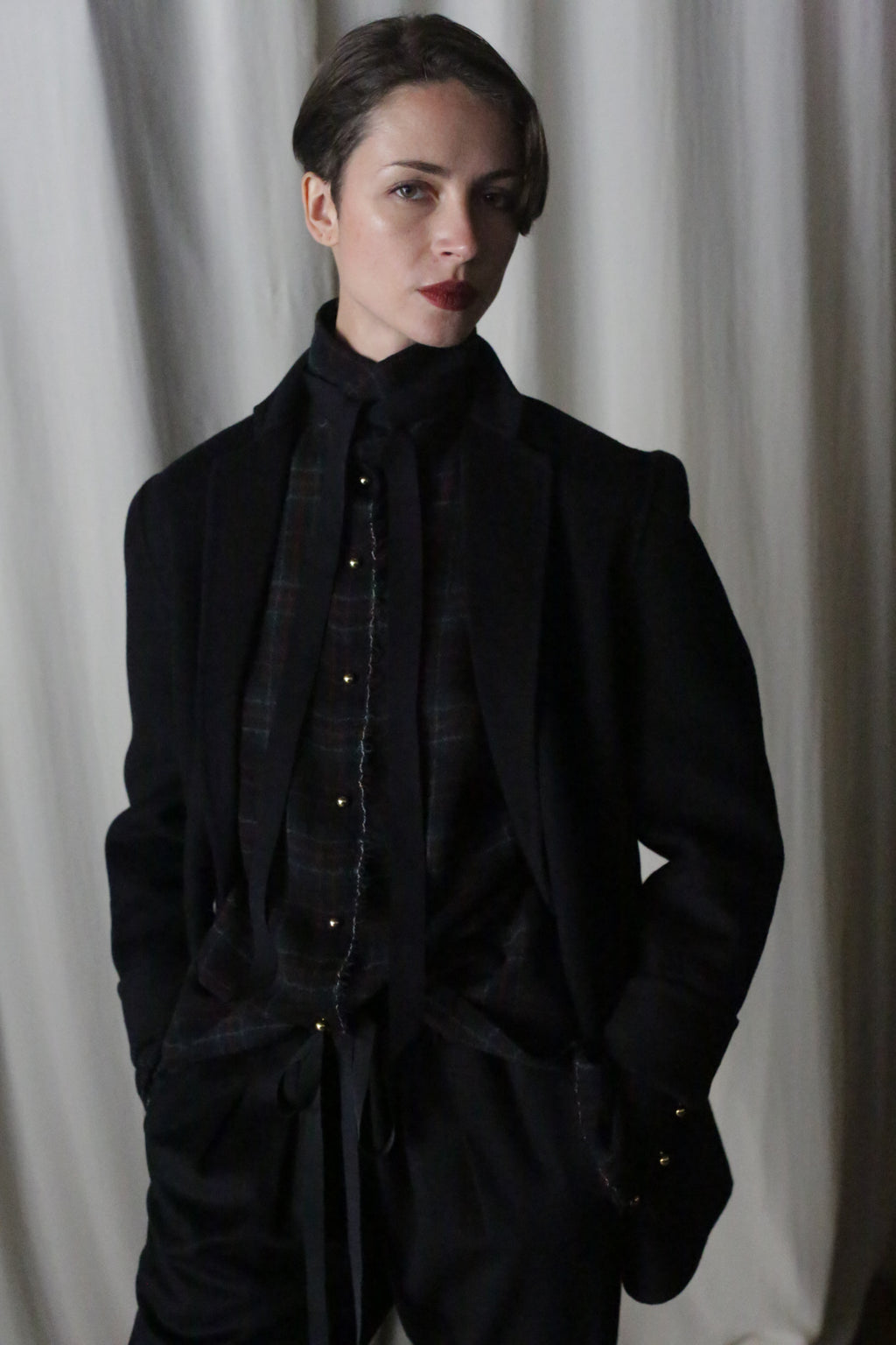 In a light-colored, draped backdrop, a short-haired individual in a dark festive plaid shirt and black suit, highlighted by the Simple Shirt in Japanese Wool Gauze Forest Plaid, stands with hands in pockets.
