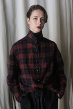 Load image into Gallery viewer, A person with short hair wears a Lan Jaenicke Simple Shirt in Japanese Wool Gauze Wine Plaid. They stand against a light curtain backdrop with hands in the pockets of dark pants, showing a neutral expression.