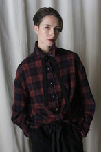 Load image into Gallery viewer, A person with short hair, dressed in a Simple Shirt | Japanese Wool Gauze Wine Plaid by Lan Jaenicke, stands against a light curtain and looks at the camera with a neutral expression.