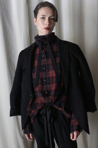 A person stands before a light curtain in a black jacket over a Simple Shirt | Japanese Wool Gauze Wine Plaid, flaunting a ruffled collar and black ribbon tie. With short hair and dark lipstick, they gaze directly at the camera.