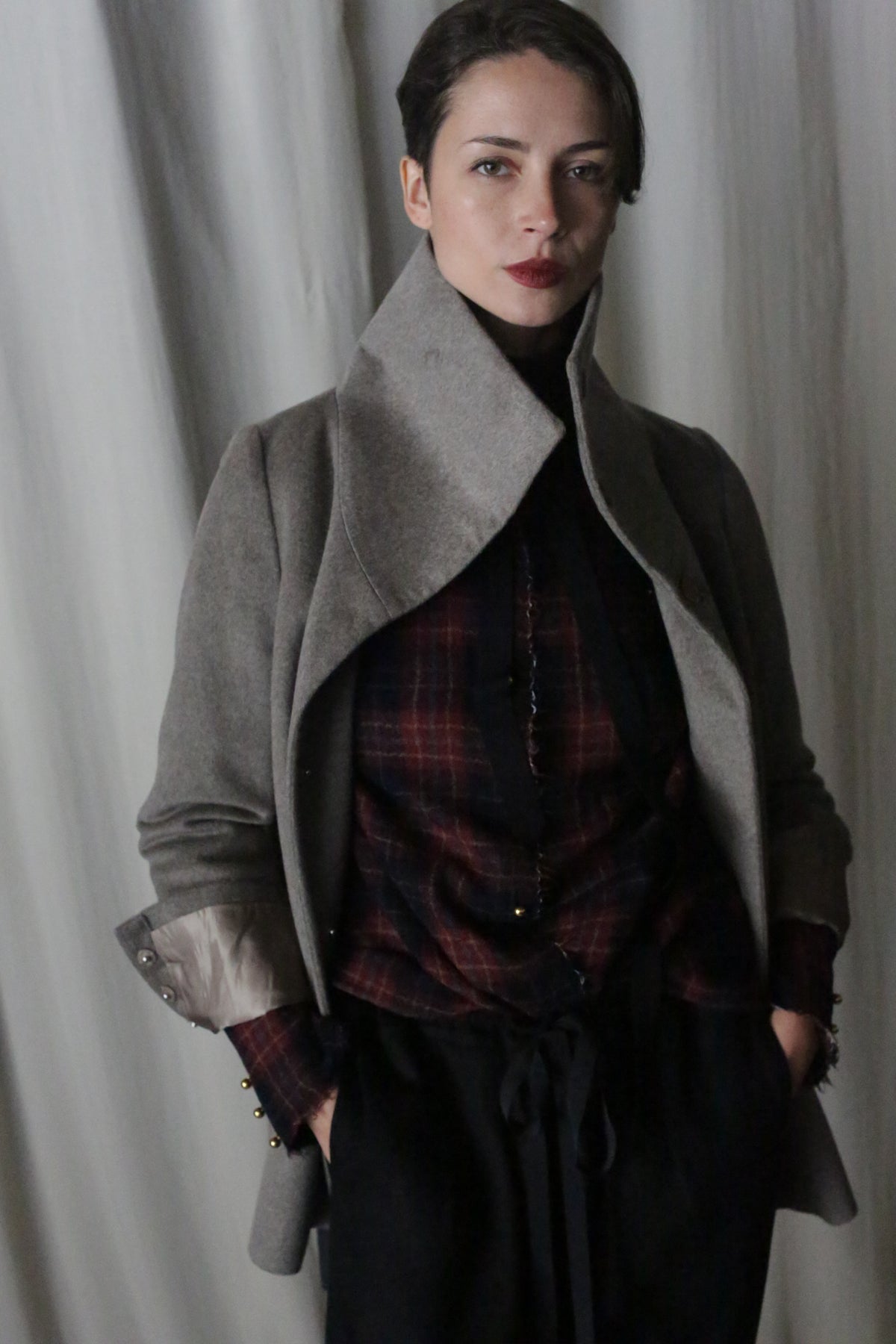 A person stands confidently against a gray curtain, wearing the Stamford II in Undyed Natural over a red plaid shirt and dark pants. They have short hair, gaze into the camera with a slight smile, and keep their hands in their pockets, enhancing their poised demeanor.
