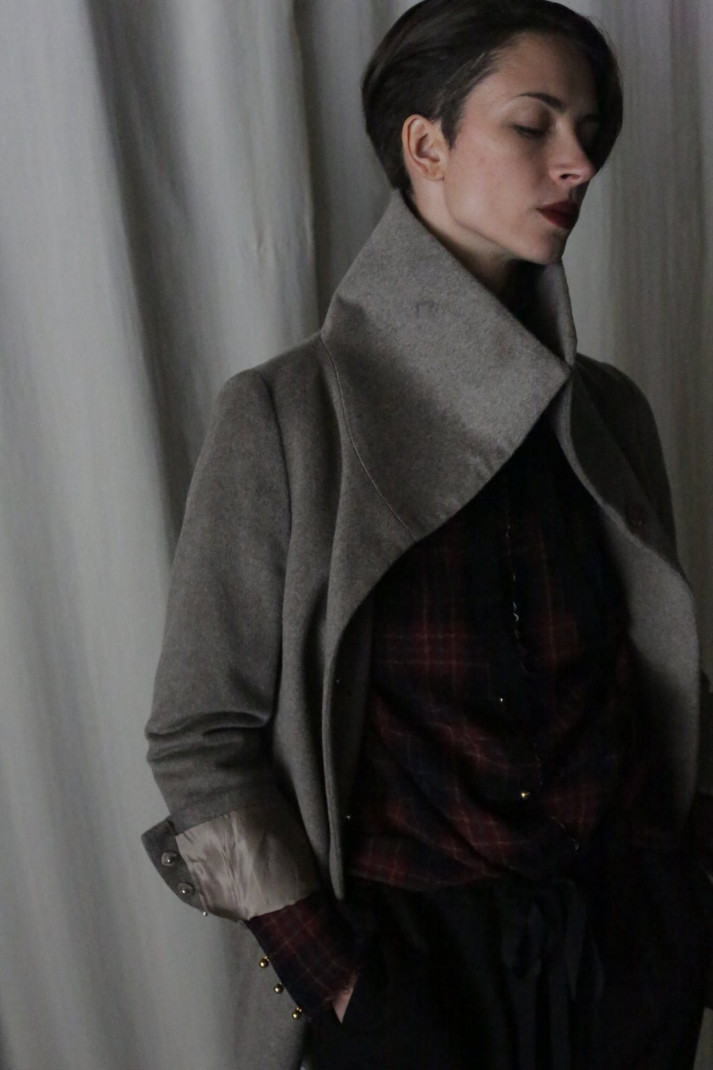 A person with short hair poses against a plain background, wearing the Stamford II—a signature jacket in undyed natural woven cashmere. Their eyes are closed and hands in pockets as a dark plaid shirt subtly complements the coat's wide collar.