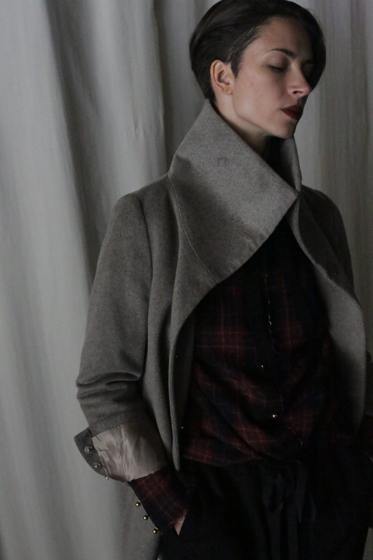 A person with short hair poses against a plain background, wearing the Stamford II—a signature jacket in undyed natural woven cashmere. Their eyes are closed and hands in pockets as a dark plaid shirt subtly complements the coat's wide collar.