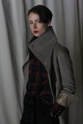 A person stands confidently in the Stamford II | Undyed Natural high-collared jacket over a plaid shirt and dark pants. They have short dark hair and red lipstick, posed against a backdrop of soft, draped fabric.