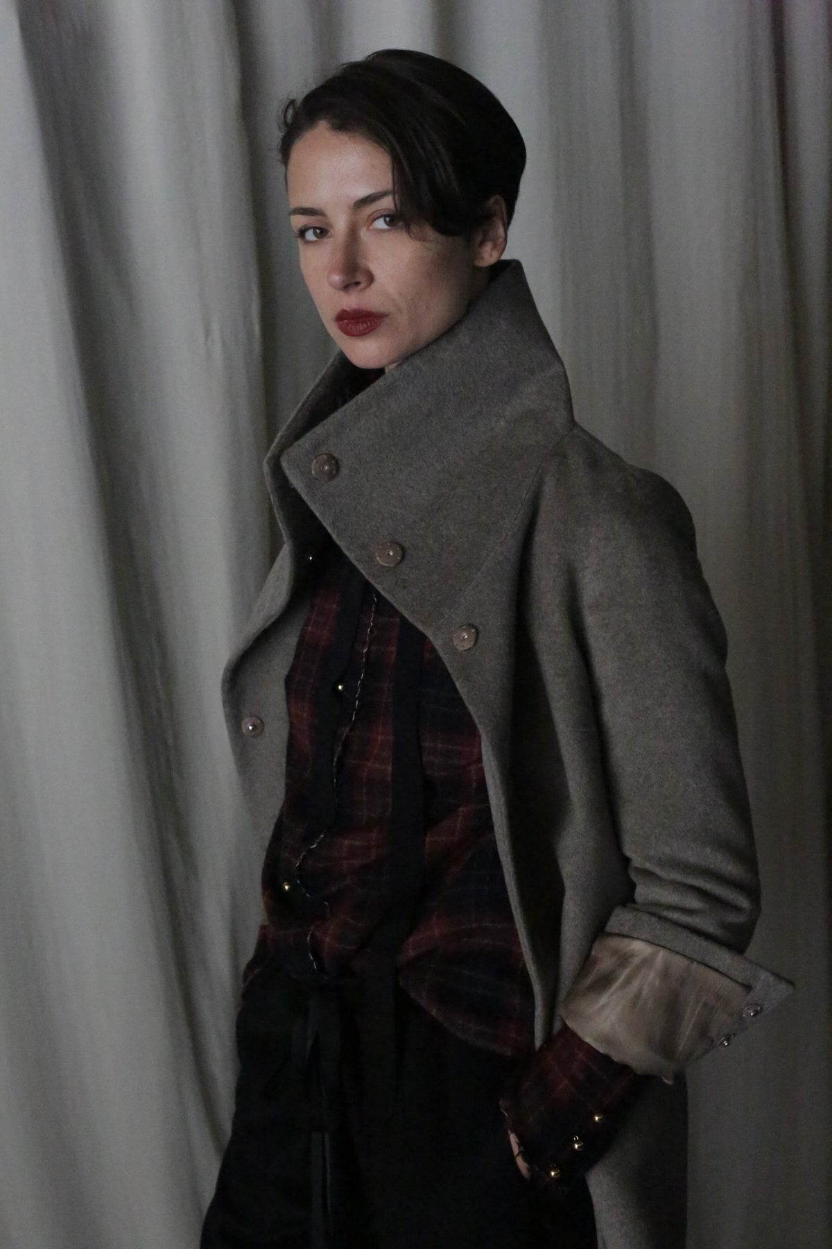 A person stands confidently in the Stamford II | Undyed Natural high-collared jacket over a plaid shirt and dark pants. They have short dark hair and red lipstick, posed against a backdrop of soft, draped fabric.