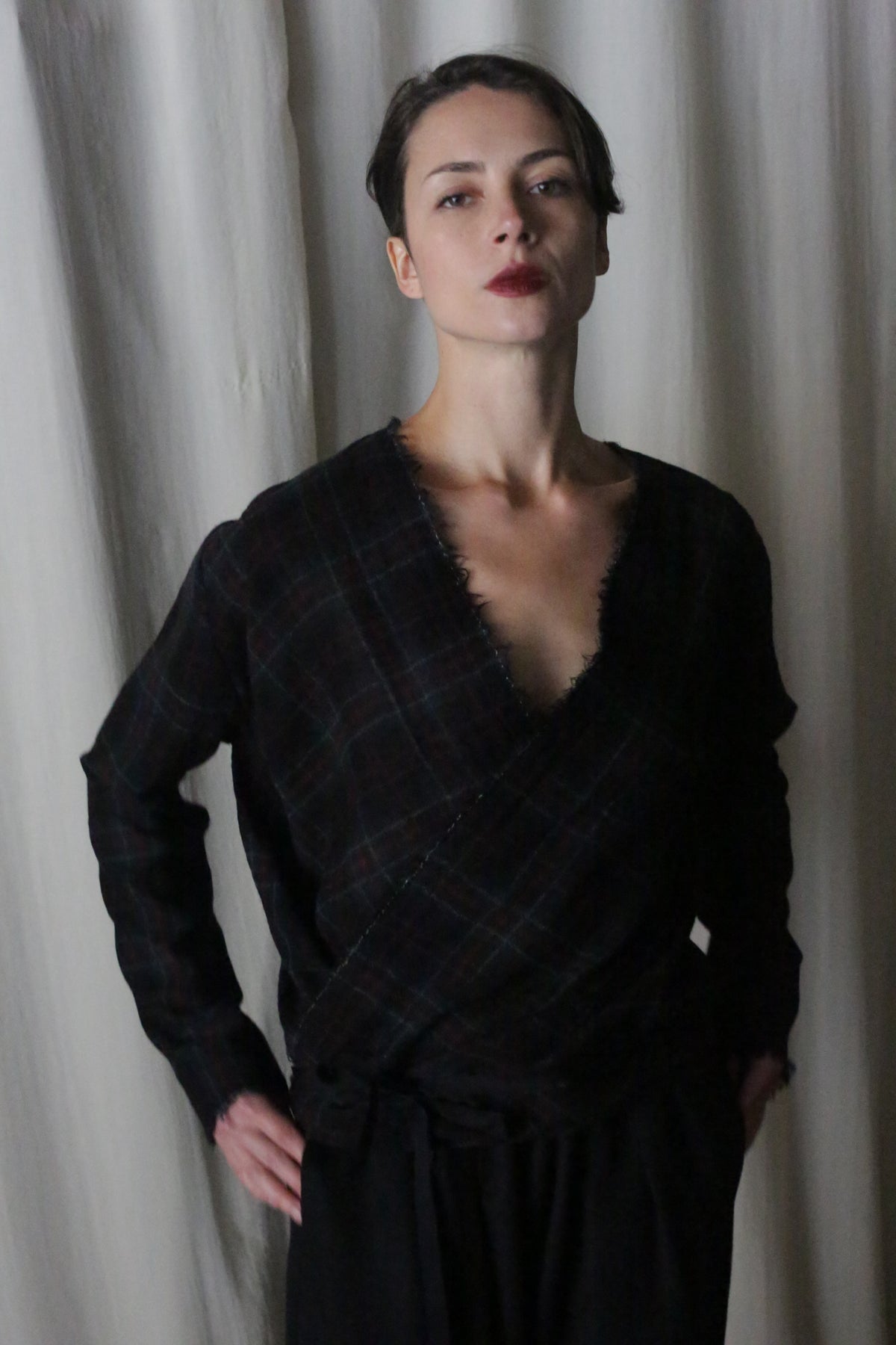 A person stands confidently in a Japanese wool gauze Forest plaid wrap top called the X Blouse with Sleeves, paired with black pants. With short dark hair and red lipstick, they stand against light-colored curtains, evoking Atelier Lan Jaenicke's refined elegance.