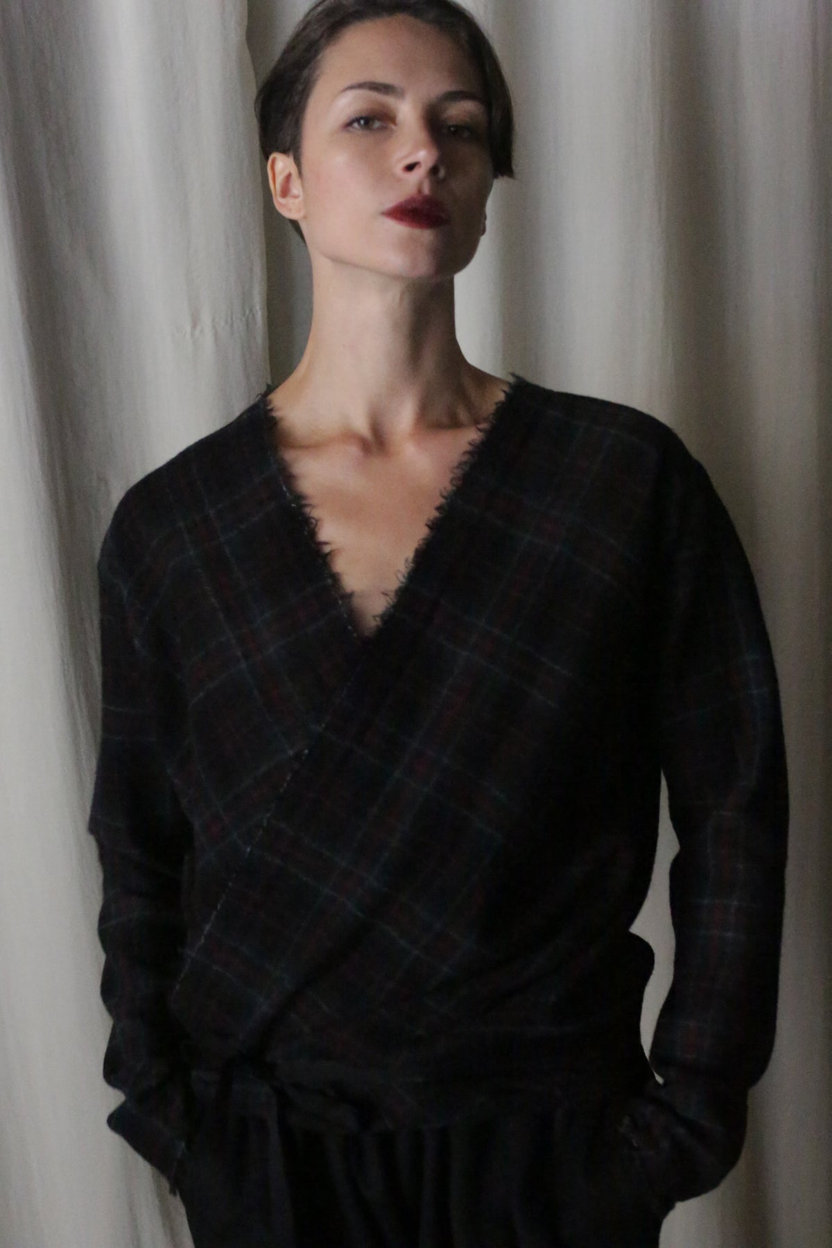 A person with short hair and red lipstick wears an Atelier Lan Jaenicke X Blouse With Sleeves in Japanese Wool Gauze Forest Plaid, standing against a light curtain backdrop.