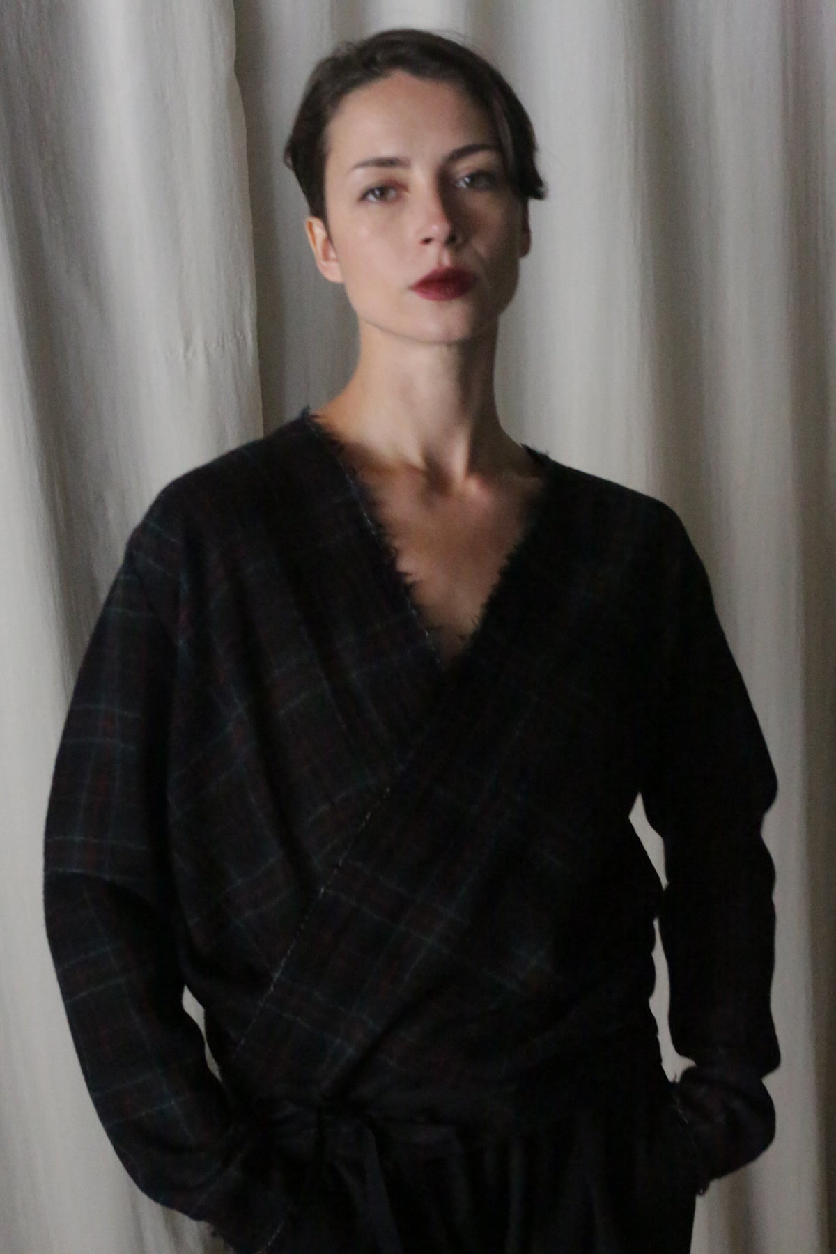 A person stands before a light curtain, wearing an Atelier Lan Jaenicke X Blouse with sleeves in Japanese Wool Gauze Forest Plaid. With short hair and a neutral expression, their hands rest in pockets as soft lighting emphasizes the blouse's elegant simplicity against the plain background.