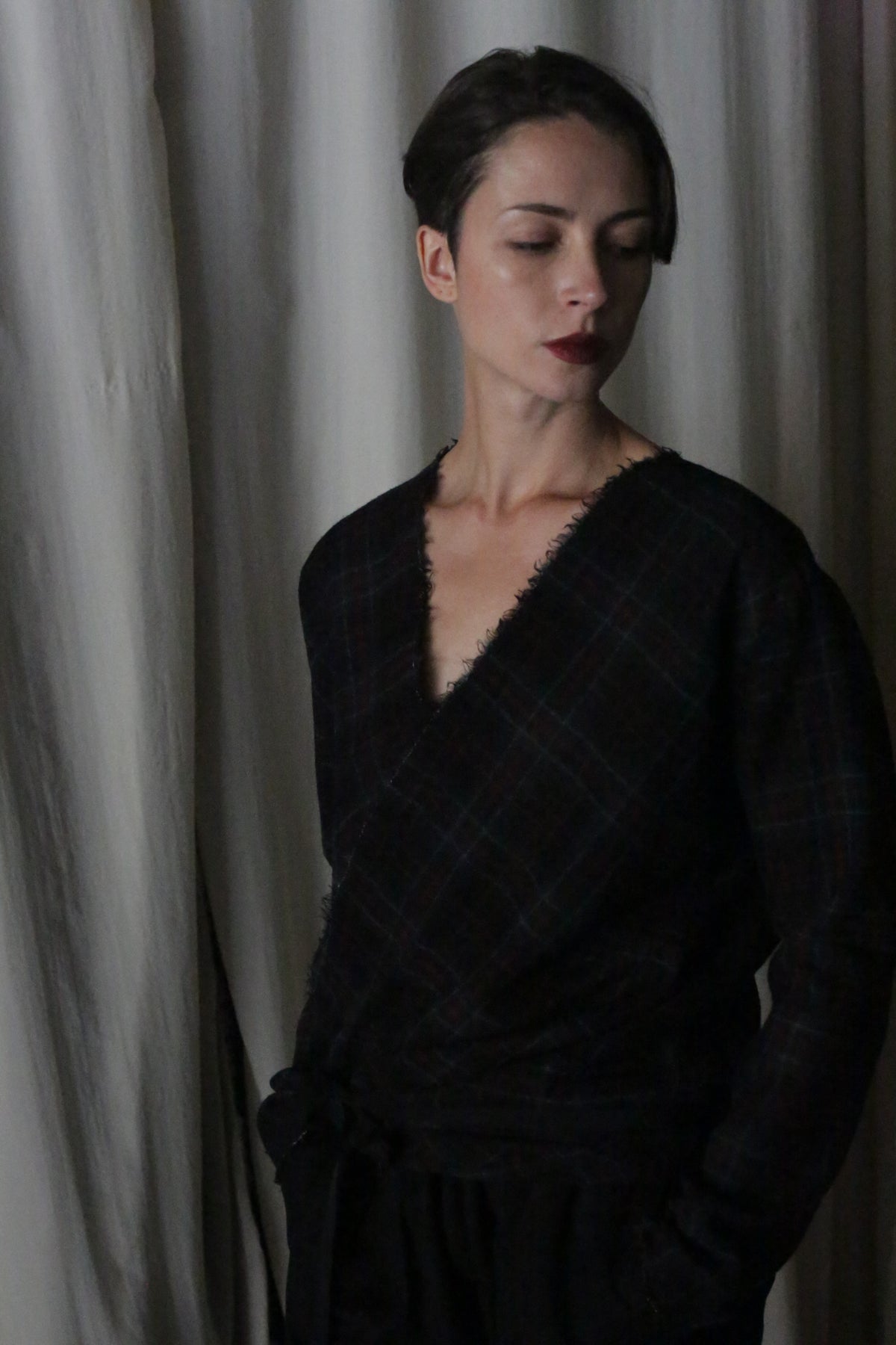 A person with short dark hair stands against draped white fabric, wearing an Atelier Lan Jaenicke X Blouse With Sleeves in Japanese wool gauze forest plaid. Their serious expression and closed eyes add depth to the scene.