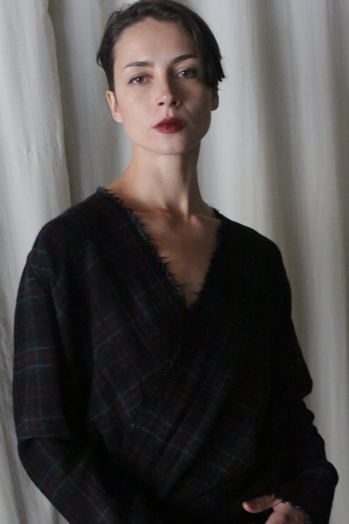 A person with short dark hair stands before a softly lit curtain, wearing an Atelier Lan Jaenicke X Blouse With Sleeves | Japanese Wool Gauze Forest Plaid. The V-neck design complements their serious expression and natural makeup accentuated by dark lipstick.