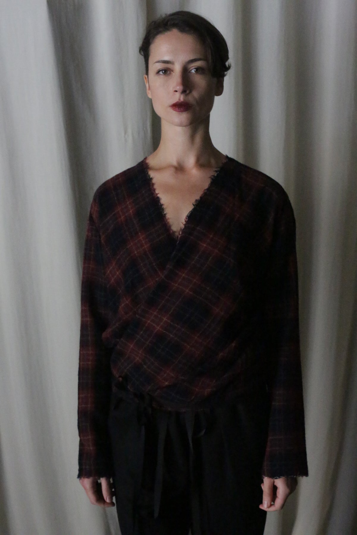 A person with short dark hair stands in front of a curtain, wearing the handmade X Blouse With Sleeves in Japanese Wool Gauze Wine Plaid by Atelier Lan Jaenicke, paired with black pants. They have a neutral expression, and the background is softly lit.