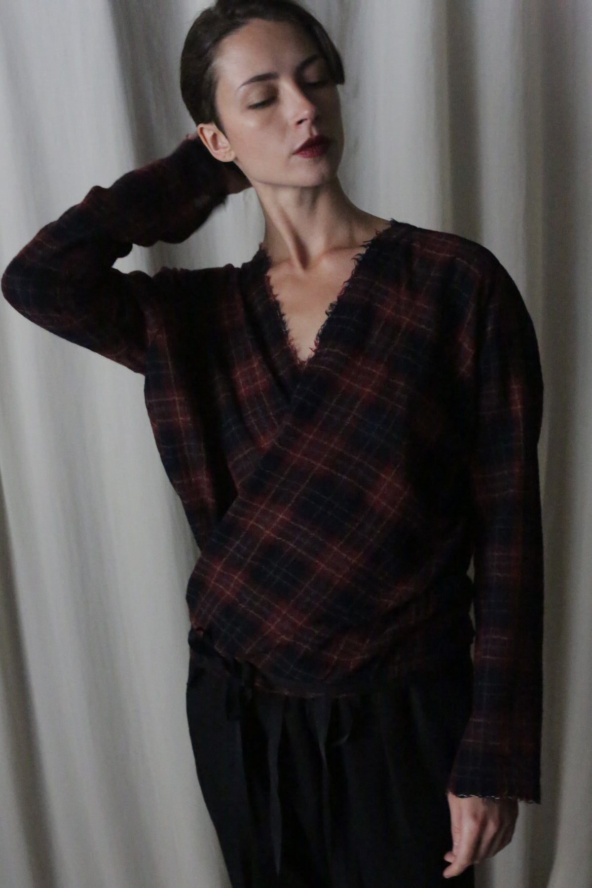 With eyes closed and head tilted slightly back, a person gracefully stands against a light curtain, resting one arm behind their head. They're clad in black pants and a dark low-waste X Blouse With Sleeves in Japanese Wool Gauze Wine Plaid.