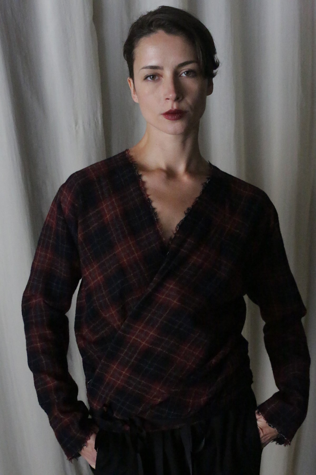 A person stands before a light curtain, wearing the X Blouse With Sleeves made from Japanese Wool Gauze in Wine Plaid, with a V-neck and long sleeves. The low-waste design, handmade at Atelier Lan Jaenicke, matches their short hair and neutral expression as they rest hands in pockets.