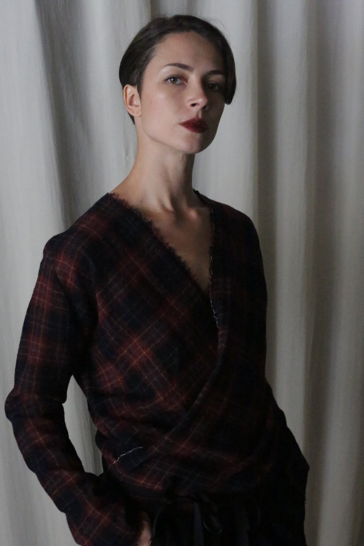 A person with short hair stands before a light curtain, wearing a handmade X Blouse With Sleeves in Japanese Wool Gauze Wine Plaid from Atelier Lan Jaenicke. They have one hand in their pocket and a serious expression.