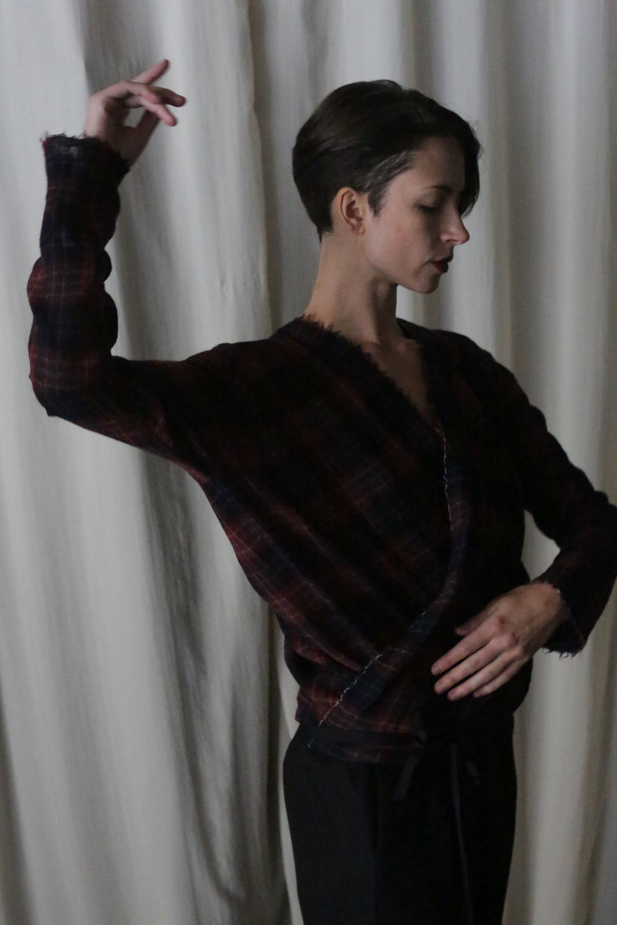 A person with short hair poses gracefully, one arm raised and the other on their waist, wearing black pants and the X Blouse With Sleeves in Japanese Wool Gauze Wine Plaid. This Atelier Lan Jaenicke piece exudes elegance as they stand before soft white curtains.