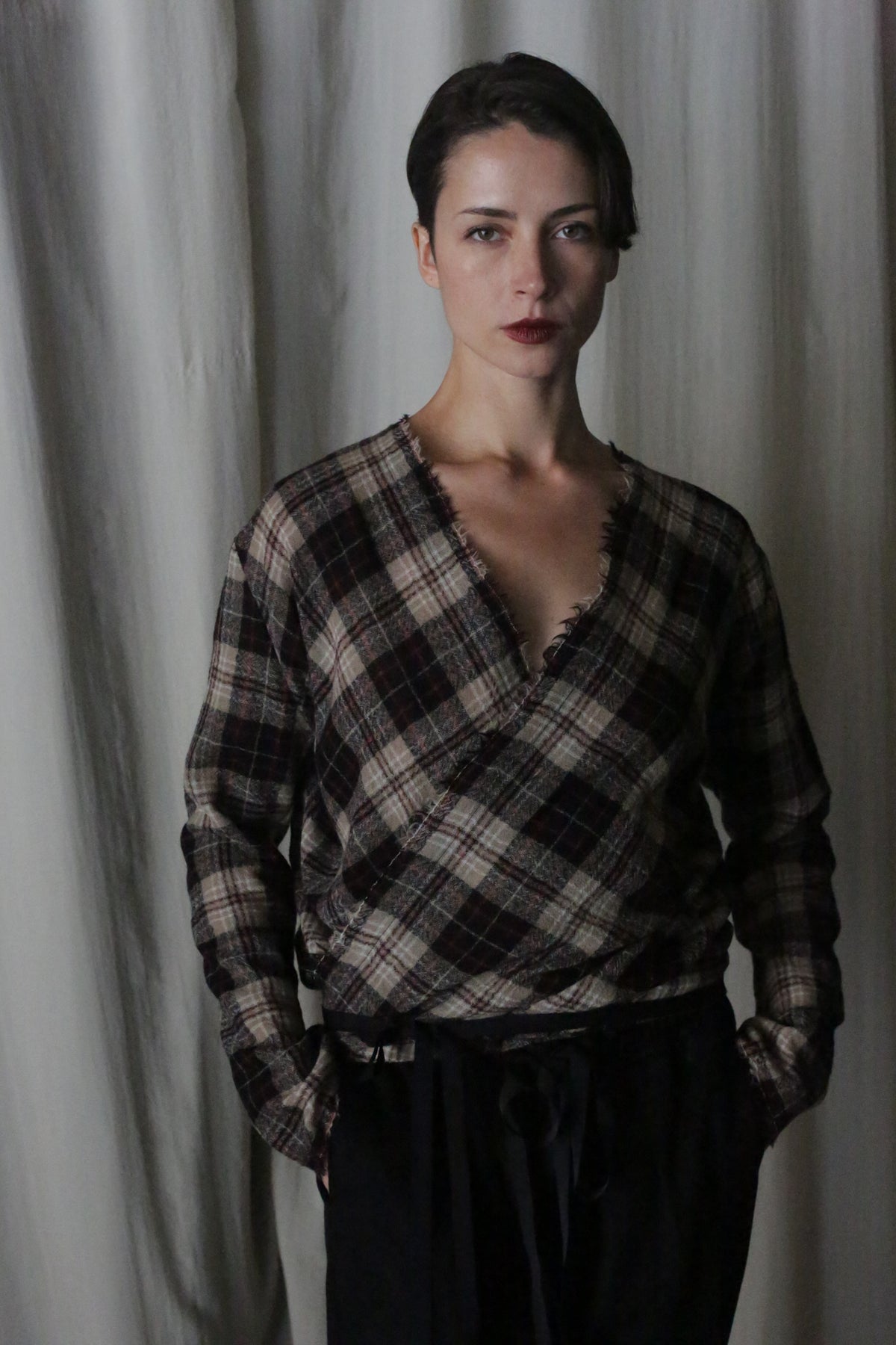 A person with short dark hair stands against a light curtain backdrop, wearing an X Blouse With Sleeves in Japanese Wool Gauze Taupe Plaid. They have their hands in their pockets and stare neutrally at the camera.