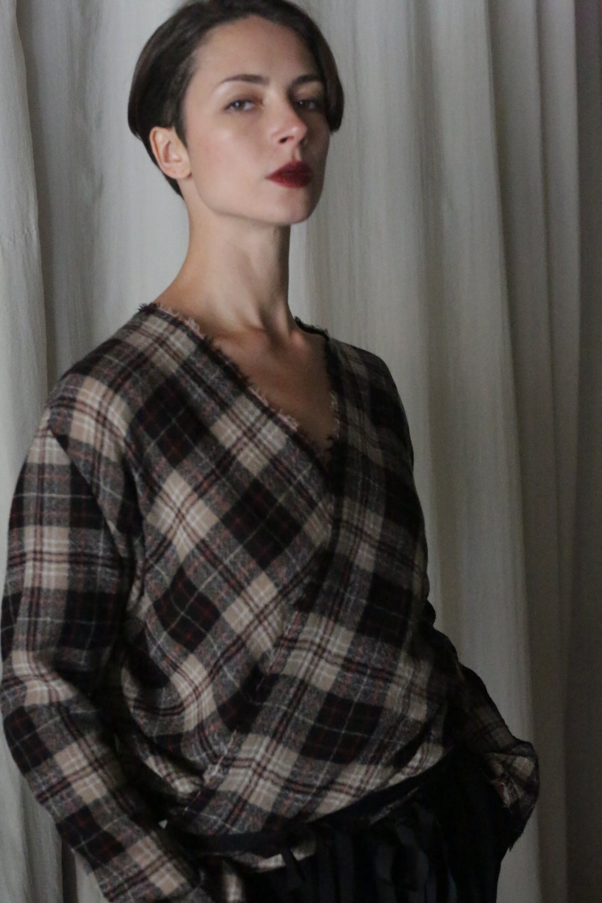 A confident individual with short hair and dark lipstick stands against light curtains, wearing the Atelier Lan Jaenicke X Blouse With Sleeves in Japanese Wool Gauze Taupe Plaid, with one hand in their pocket.