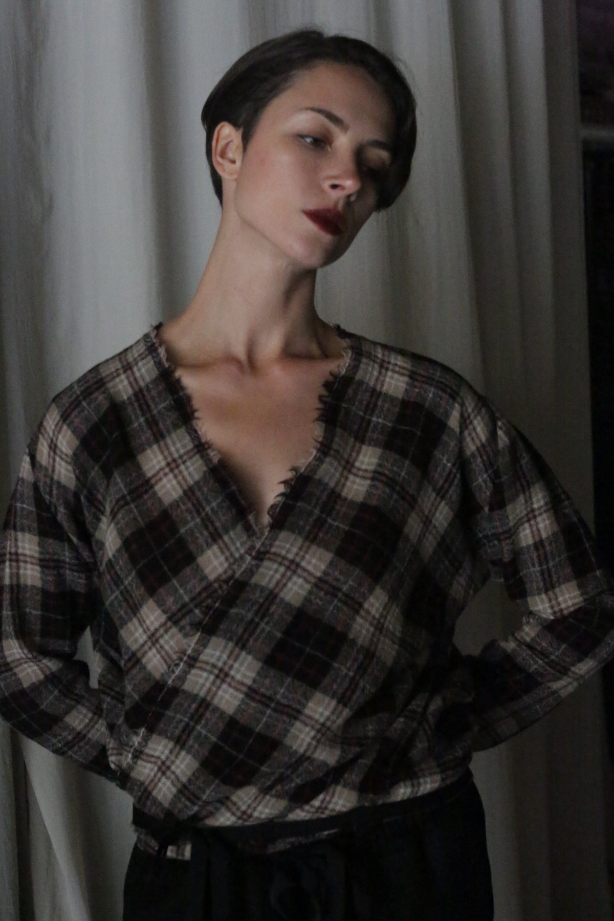 In front of a curtain backdrop, a person with short hair poses thoughtfully, wearing the X Blouse With Sleeves in Japanese Wool Gauze Taupe Plaid from Atelier Lan Jaenicke. With a slight head tilt and dark lipstick, they exude refined elegance.