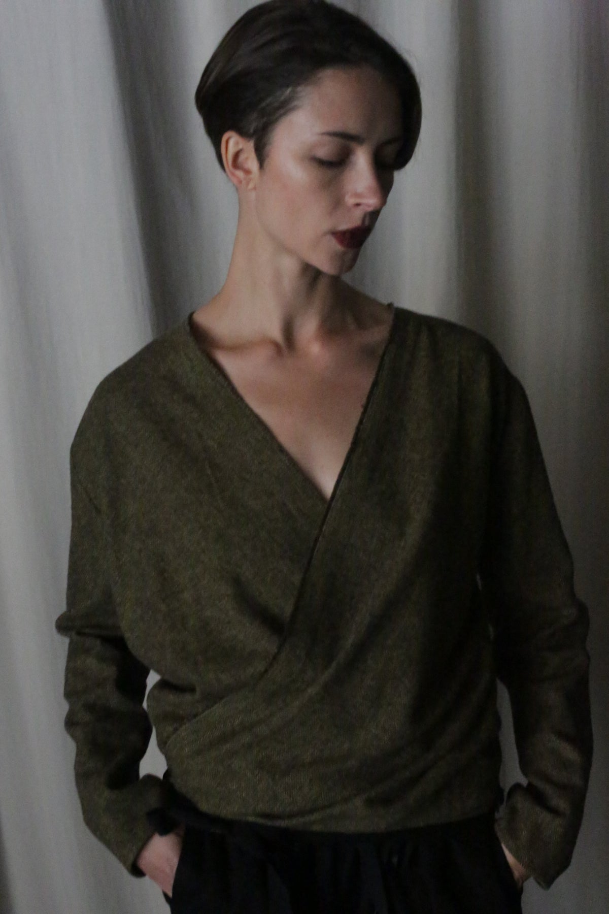 A person with short hair stands against a curtain in a dark green X Blouse With Sleeves made of brushed cotton moss herringbone, paired with dark pants. They have their hands in their pockets and gaze thoughtfully downwards, soft lighting casting shadows on this versatile wardrobe staple.