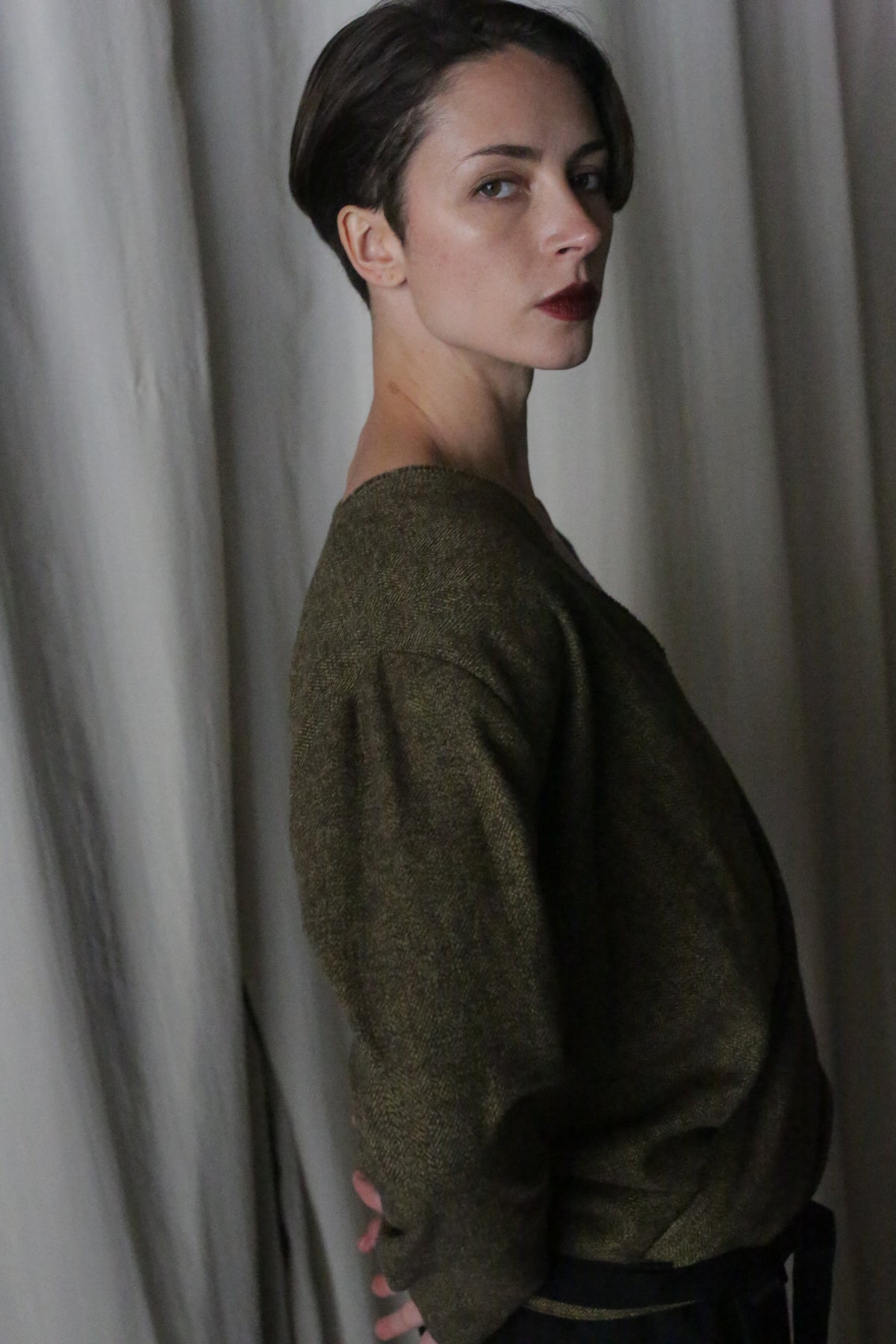 A person with short hair, wearing a dark brushed cotton moss herringbone X Blouse with sleeves and dark pants, poses sideways against light-colored curtains, looking toward the camera with a serious expression.